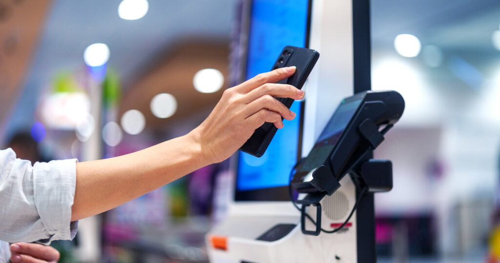 Person nutzt ein Smartphone, um an einem Selbstbedienungsterminal zu bezahlen, im Hintergrund unscharfe Lichter eines modernen Einkaufszentrums.