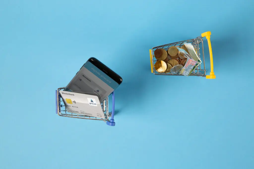 Zwei Einkaufswagen auf einem blauen Hintergrund, einer mit einem Smartphone und einer Bankkarte, der andere gefüllt mit Münzen und Geldscheinen, symbolisieren digitale und traditionelle Zahlungsmethoden.