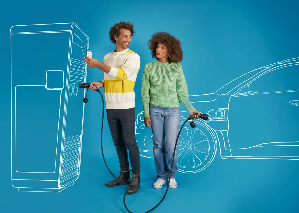 Paar steht vor einer Ladesäule für Elektroautos und hält Ladeanschlüsse in der Hand. Der Mann zeigt auf ein Smartphone, während die Frau interessiert zusieht. Im Hintergrund ist eine stilisierte Skizze eines Elektrofahrzeugs auf blauem Hintergrund zu sehen.