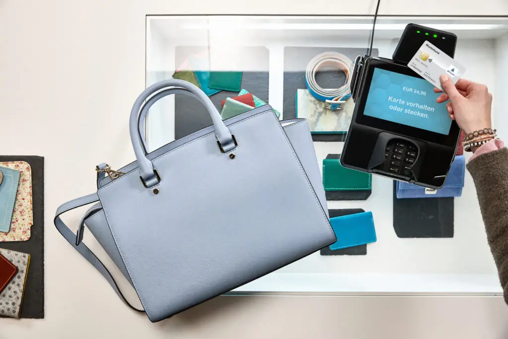 Moderne hellblaue Handtasche neben einem Kartenlesegerät, während eine Person ihre Kreditkarte für eine Zahlung vorhält. Im Hintergrund sind verschiedene Geldbörsen und Accessoires in einem Schaufenster sichtbar.