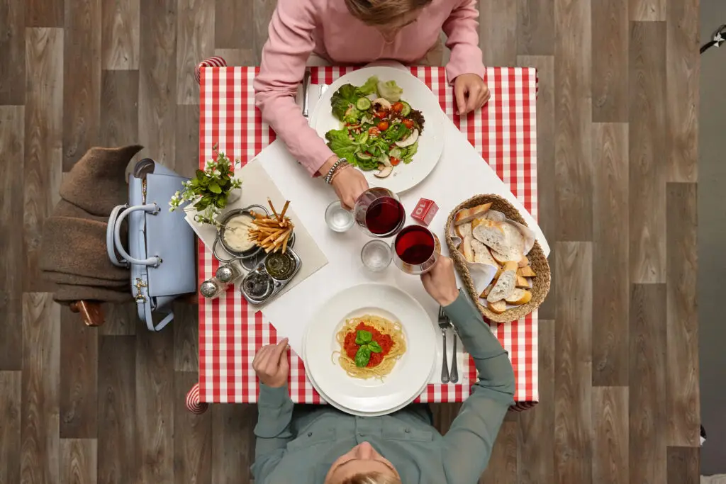 Tischdecke im karierten Design mit zwei Personen, die ein köstliches Essen genießen; im Vordergrund Spaghetti mit Tomatensauce und Basilikum, im Hintergrund ein frischer Salat, dazu Getränke und Brot. Ideal für ein gemütliches Restaurantambiente.