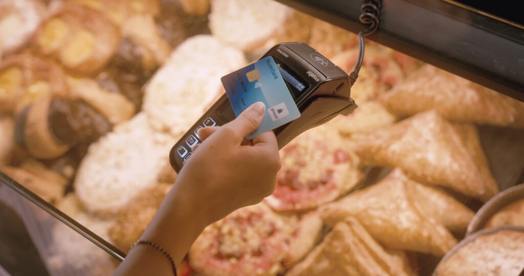 A person uses a contactless payment terminal to make a payment with a bank card for baked goods in a display. Various baked goods are visible in the background.