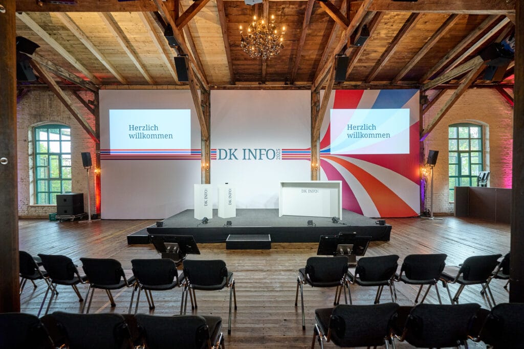 Veranstaltungsraum für DK INFO 2025 mit stilvoller Dekoration, geeigneten Sitzplätzen und Präsentationsflächen, die einen einladenden Eindruck vermitteln. Die Bühne ist für eine Begrüßungsansprache vorbereitet, mit digitalen Displays und moderner Technik.
