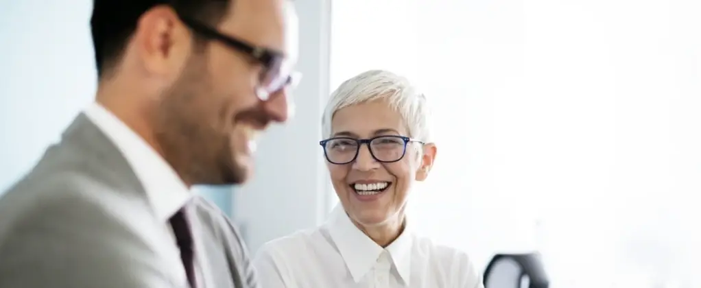 Lächelnde Geschäftspartner in einem modernen Büro, die sich unterhalten und eine positive Atmosphäre ausstrahlen. Die Frau trägt eine weiße Bluse und Brille, während der Mann in einem grauen Anzug gekleidet ist.