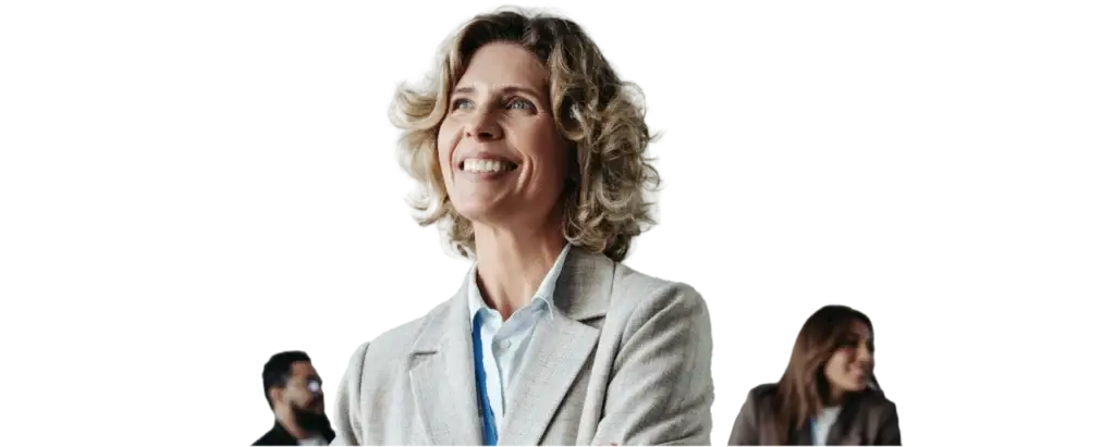 Woman with curly hair and a smiling expression standing in a modern office environment. Two colleagues can be seen in the background, engrossed in a conversation. The scene conveys a sense of teamwork and a positive working atmosphere.