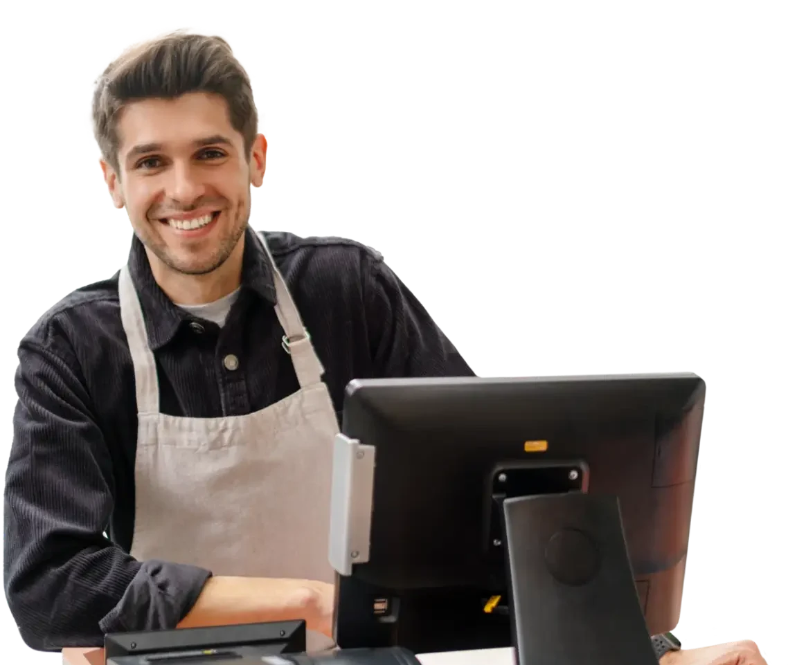 Lächelnder junger Mann in einem schwarzen Hemd und einer Schürze, der an einem Computerarbeitsplatz steht und freundlich in die Kamera blickt. Der Hintergrund ist neutral gehalten, um den Fokus auf den Mitarbeiter zu lenken.