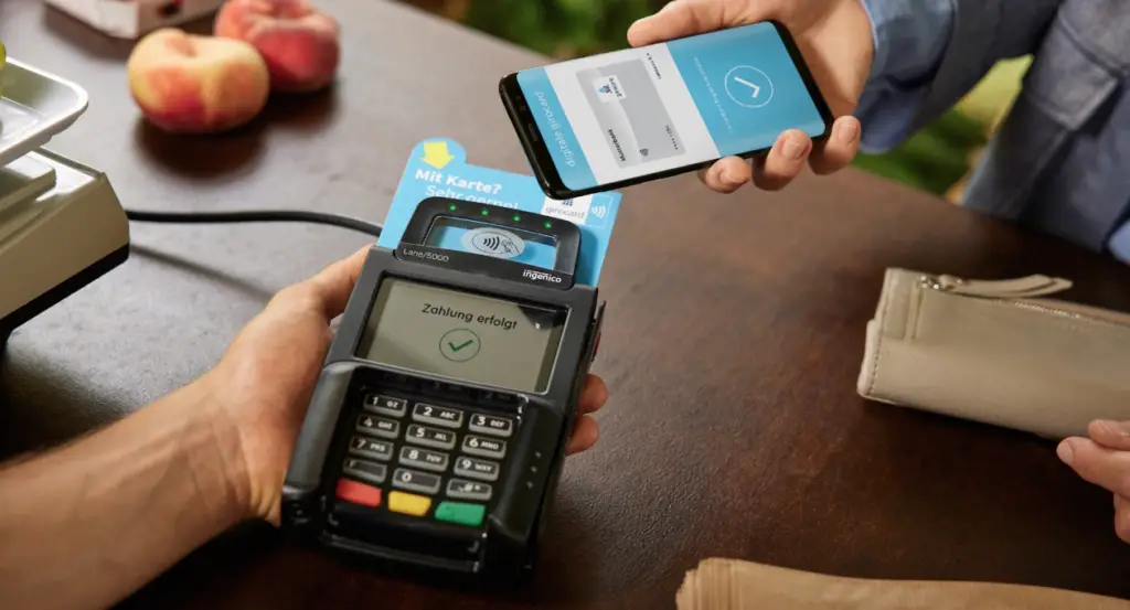 Mobile Zahlung mit Smartphone an einem Kartenlesegerät, das den erfolgreichen Abschluss der Transaktion anzeigt. Im Hintergrund sind frische Äpfel und eine Geldbörse zu sehen.
