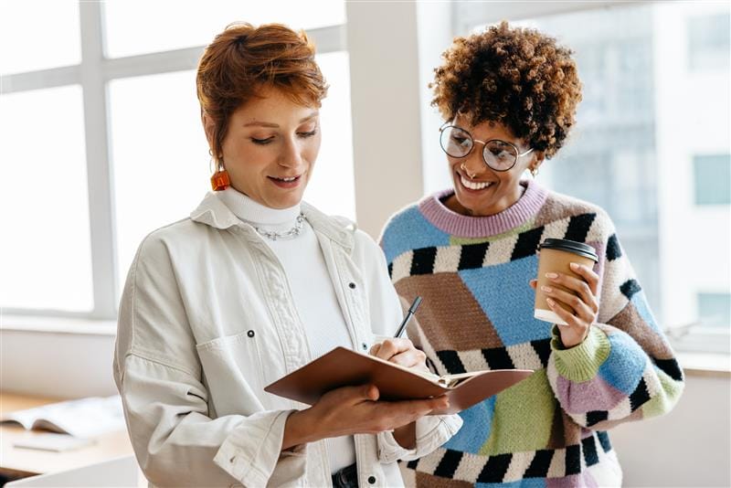 Two women are standing in a bright room talking. One woman with short red hair is holding a notebook and writing, while the other woman with curly hair is holding a coffee mug and smiling. Both are wearing fashionable, casual clothes and appear to be working on a creative project.