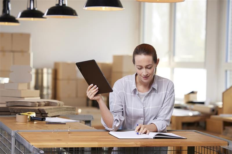 Woman in a modern office, taking notes in a notebook with a tablet in her hand, surrounded by boxes and office supplies. Ideal for topics such as productivity, office organization and digital work.