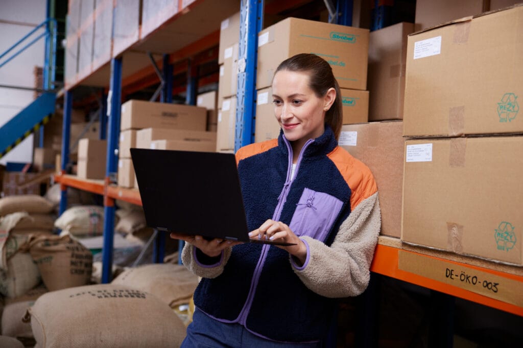 A woman in a warehouse is working on a laptop, surrounded by boxes and bags. She wears a warm jacket and smiles as she looks at the screen. The environment shows organized warehousing and modern logistics.