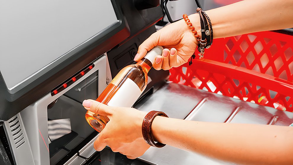 A person scans a bottle at a self-service checkout machine in a supermarket, while the shopping cart is visible in the background.