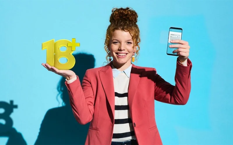 Young woman with curly hair holding a yellow 18+ sign in one hand and a smartphone in the other, against a light blue background. She is wearing a red blazer and a striped top and is smiling.