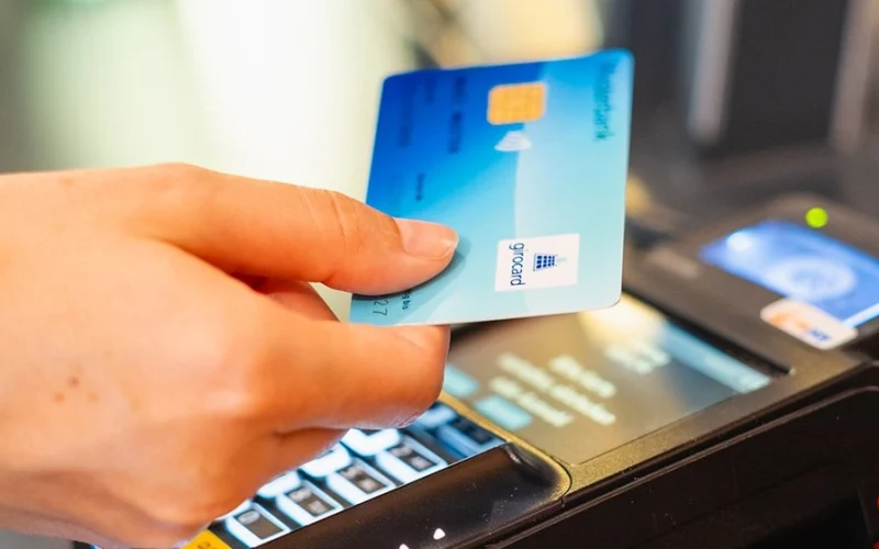 Hand holding a credit card at a card reader while a transaction is being carried out. The card a modern design and is suitable for contactless payment.