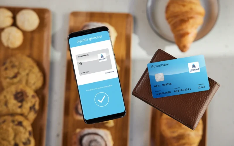 Smartphone with digital Girocard and a physical Girocard in a wallet, surrounded by cookies on a wooden board. The picture illustrates the use of modern payment methods and the integration of digital financial solutions in everyday life.