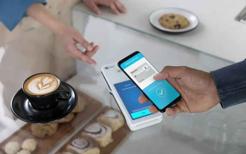 A person uses a smartphone to pay in a café while a cup of coffee and various baked goods are on the table. The smartphone screen displays a digital voucher card that is used for authentication.