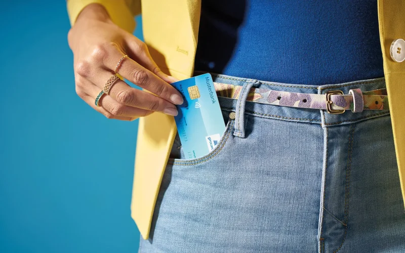 Lady a hand putting a bank card in the pocket of a pair of jeans while wearing a yellow blazer jacket, against a blue background.