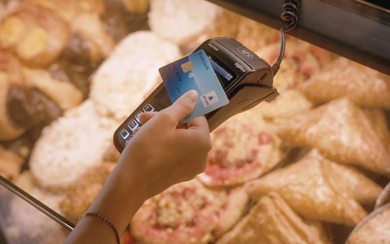 A person uses a contactless payment terminal to make a payment with a bank card for baked goods in a display. Various baked goods are visible in the background.