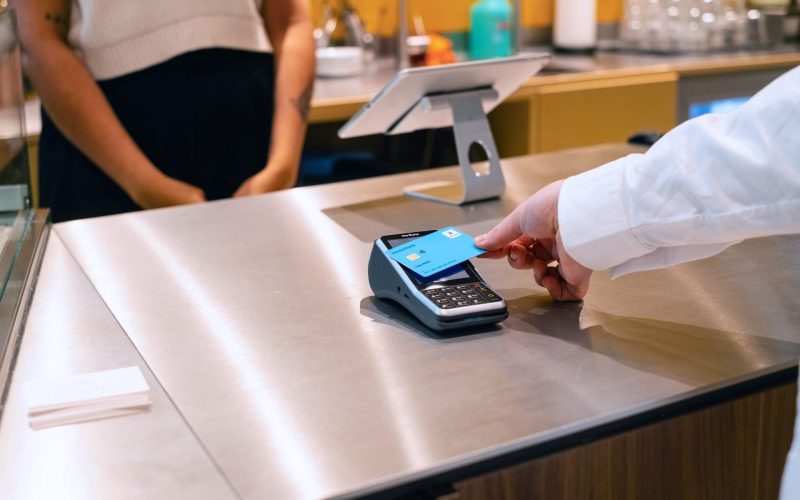 Payment process at the checkout with a card reader in a modern café, while a person holds a credit card in front of the device. Another person can be seen in the background waiting for the payment process.
