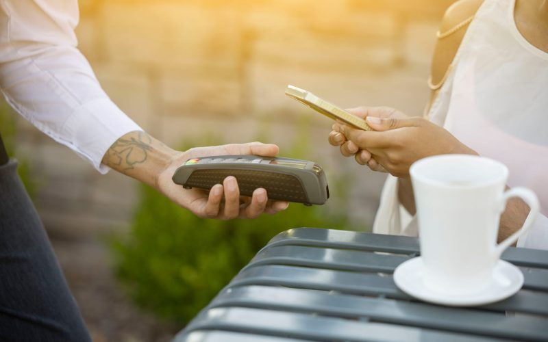 Payment process in a café, where a customer uses his smartphone to pay while an employee hands over the card reader. A cup of coffee is visible on the table in the background.