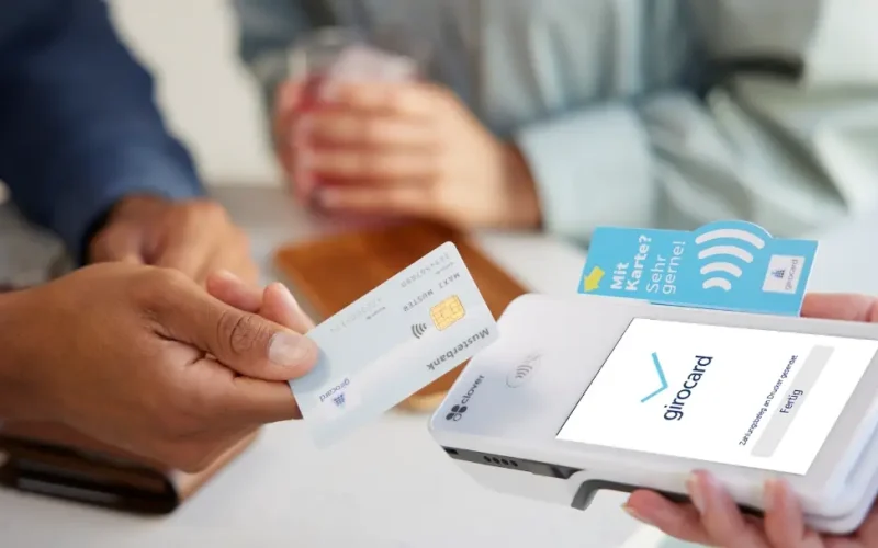Two people make a contactless payment while one hand holds a girocard to a payment terminal. A glass and a wallet are visible in the background, conveying a relaxed café situation.