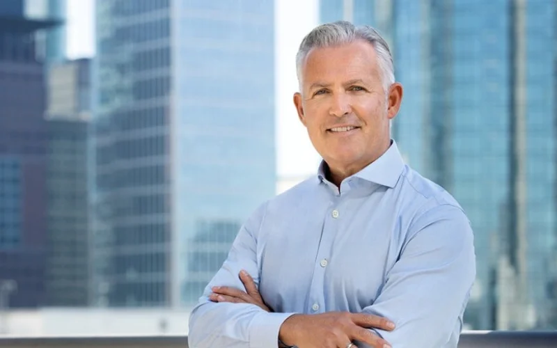 Businessman with gray hair standing in front of skyscrapers in a modern urban environment, smiling while crossing his arms.