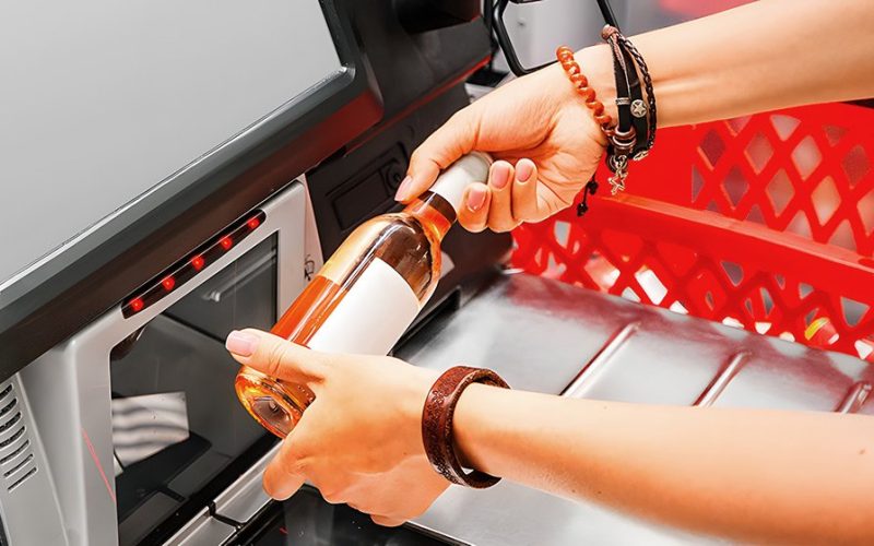 A person scans a bottle at a self-service checkout machine in a supermarket, while the shopping cart is visible in the background.