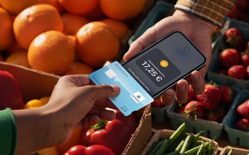 Payment with smartphone and credit card at a market surrounded by fresh fruit and vegetables, including oranges, peppers and strawberries. The amount of €17.25 is displayed.