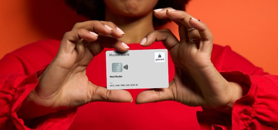 Frau in rotem Oberteil hält eine moderne Bankkarte der Musterbank in den Händen, auf einem orangefarbenen Hintergrund. Die Karte zeigt ein kontaktloses Zahlungssymbol und den Namen Maxi Muster.