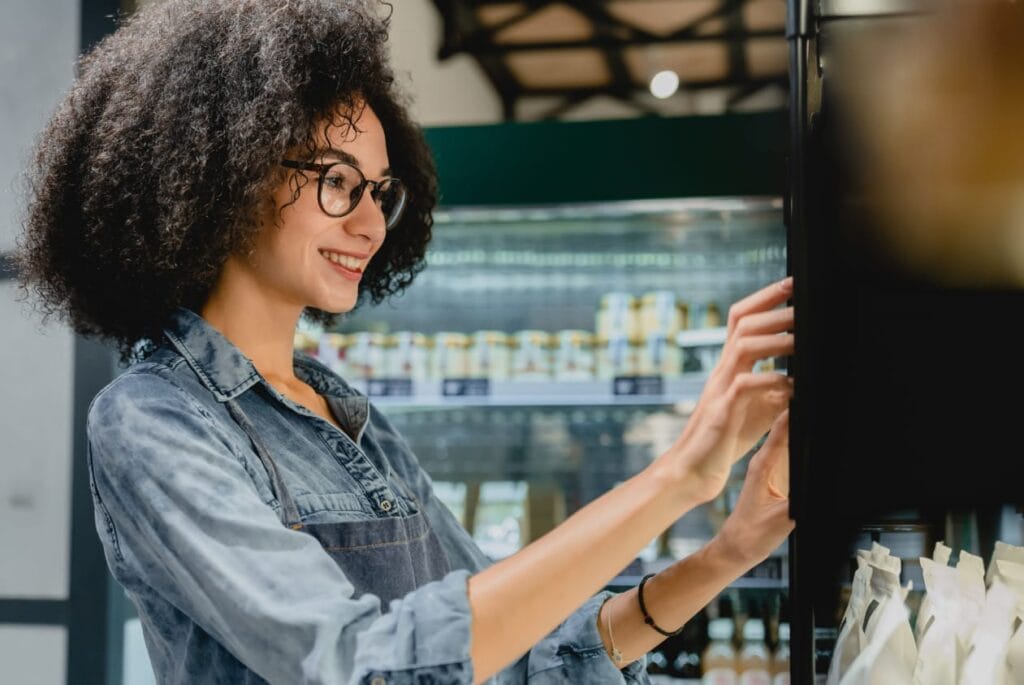 Young woman with curly hair and glasses looking through shelves of products in a modern store, smiling and interested.