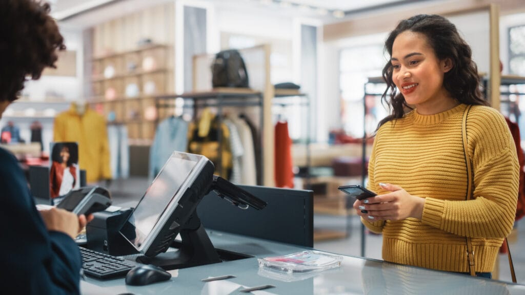 Woman in a fashion store at the checkout, smiling with a smartphone in her hand while the cashier carries out a payment transaction. Various items of clothing and accessories are visible in the background.