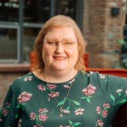 Portrait of a smiling woman with blonde hair and glasses wearing a green floral pattern. The background shows a modern interior design with windows and plants.