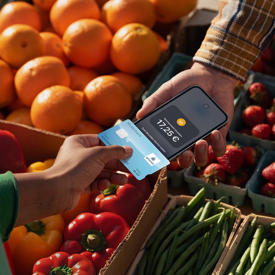 Payment with smartphone and credit card at a market surrounded by fresh fruit and vegetables, including oranges, peppers and strawberries. The amount of €17.25 is displayed.